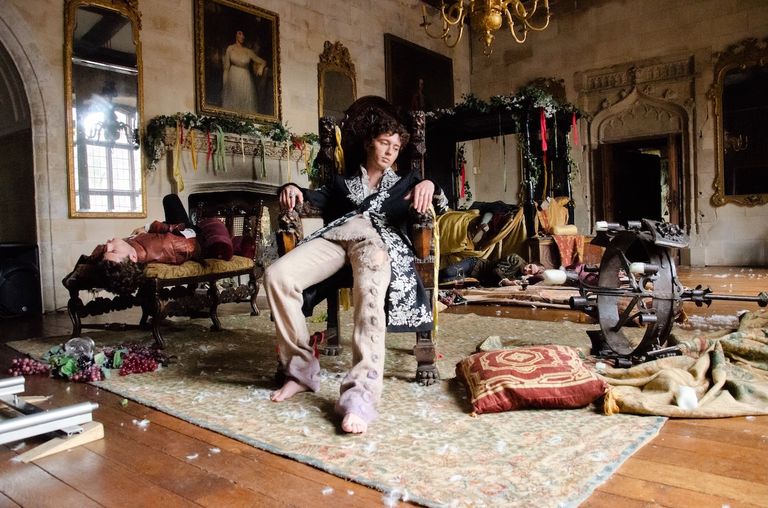 Pop singer Troye Sivan lounging on an ornate wooden throne in an elaborate, disorderly vintage room with classic paintings.